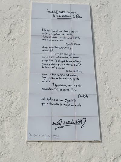 This media file is a photograph of a ceramic tile plaque mounted on a textured white wall. The plaque displays a handwritten poem in Spanish, with some words written in a larger, bolder script.

The poem's title is "PALABRAS PARA COLGAR DE UNA VENTANA DE ROTA," which translates to "Words to Hang from a Window in Rota." This suggests the poem is likely located in Rota, Spain, and offers a personal perspective from a window overlooking the sea.

The poem itself speaks of the sea, foam, shore, and the horizon. It describes the sensory experience of being by the sea, mentioning sand, summer, and the sun. It also touches upon themes of childhood memories and reflection, with lines about a child, a sleepless night, and looking out from a window. The final lines reiterate the connection to the sea and seagulls.

Below the poem, there is a signature or artist's name, "ANGEL GARCIA LOPEZ," followed by a reference to "MESTER ANDALUSI" and the year "1978."

The overall impression is one of a contemplative and evocative piece of art, likely an outdoor installation, celebrating the beauty and inspiration of the sea and the specific location of Rota. The light in the image suggests it was taken during the daytime, with no specific weather conditions immediately apparent.