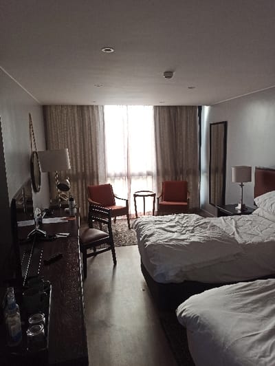 The bedroom features two beds with white linens. Two orange chairs flank a window covered by neutral-toned drapes. A desk, stocked with a glass tray, sits in the foreground. The desk's surface has miscellaneous items and a reflective quality. A wall lamp sits near the beds. The room's color palette is neutral.