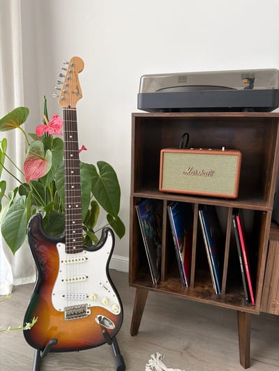 The image displays a collection of musical instruments and related paraphernalia. A sunburst Fender Stratocaster electric guitar rests on a stand, partially obscured by lush green foliage and pink anthurium flowers. Behind the guitar, a dark wood record cabinet holds a selection of vinyl records. A small Marshall speaker is placed within one of the cabinet's shelves. To the left of the cabinet, a turntable is visible, suggesting a listening area. The overall scene is one of a music enthusiast's curated space, with a warm, inviting aesthetic. The lighting suggests daytime. There is text visible on the speaker and the guitar. The text on the speaker reads "Marshall." The text on the guitar's headstock reads "Fender Stratocaster."