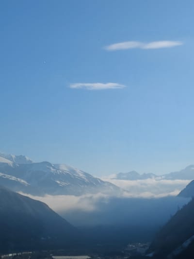 The image depicts a mountain landscape in Almasiani, Georgia. The scene is dominated by snow-capped mountains partially obscured by a low-lying fog or cloud cover in the valley. The sky is clear blue with a few thin, elongated clouds. The foreground shows the lower slopes of the mountains, descending into a valley. A small, faintly visible object is present in the upper left part of the sky. A town is also faintly visible within the valley.