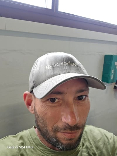 A close-up photograph features a man with a beard wearing a grey cap with the word "Flyboarders" embroidered on it. He is partially facing the camera with a slight head tilt. The background includes a glimpse of a window frame and part of a wall. A small green box with white lettering is also visible. The photo includes "Galaxy S24 Ultra" text on the upper left. There is no visible information to confirm the city of Elsinore, Denmark.