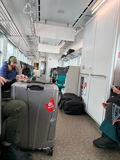 Inside a train car, passengers sit with luggage. Overhead compartments line one side of the train. A large, gray suitcase with a red "Heavy" sticker occupies a significant portion of the aisle. Other luggage pieces, including a black backpack, are nearby. People are seated, with one person wearing headphones and another appearing to use a phone. The train car appears to be modern, with teal and gray seats. Exterior snow is visible through the windows.