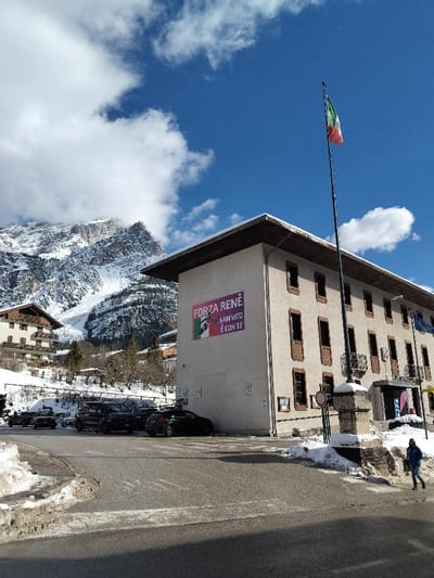 The image depicts a winter scene in a mountainous region, specifically in Serdes, Italy. A multi-story, light-colored building with dark-framed rectangular windows occupies the right side of the frame. An Italian flag flies from a tall pole in front of this building. On the building's left facade, a large banner displays "FORZA RENÉ" above an Italian flag graphic, with the text "SAN VITO È CON TE" below. The ground in front of the building consists of a partially snow-covered paved area functioning as a parking lot, where several dark-colored vehicles, including SUVs and sedans, are parked. A single individual, wearing a blue jacket, walks on a snow-lined sidewalk on the far right. In the background, prominent snow-capped mountains rise under a clear blue sky dotted with white clouds. Other chalet-style buildings are visible on a snow-covered hillside to the left of the main building. Patches of snow and bare ground are present on the roads and surrounding areas.