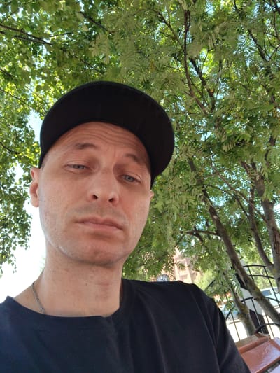 This is a selfie of a man in his late 30s to early 40s, wearing a black t-shirt and a black baseball cap. He has fair skin, short brown hair, and light blue eyes. He is looking directly at the camera with a neutral expression. The man is outdoors, with lush green trees and branches forming the background. Sunlight filters through the leaves, suggesting it is daytime. The setting appears to be a park or a garden, possibly in Mokshan, Russia, given the context. The man is wearing a silver chain necklace. The image is taken from a low angle, looking up towards the man and the trees.