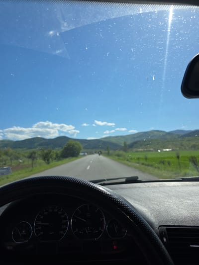 The image is taken from inside a car driving on a road in the mountains. The car's dashboard and windshield are visible. The sky is blue with some clouds. The road is straight and goes through a green valley. The speedometer shows the car is moving.
The location is not discernible as the information given is insufficient to determine it.