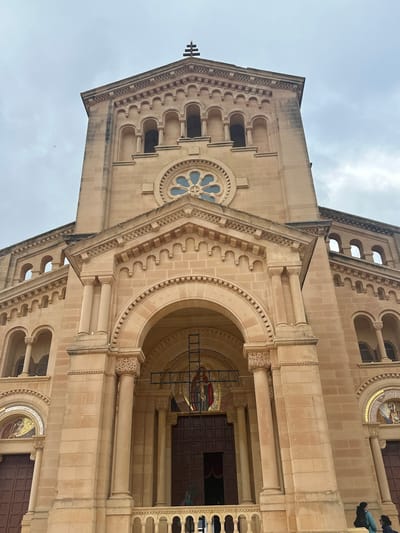 An exterior, low-angle photograph depicts the facade of the Sanctuary of Our Lady of Ta' Pinu in Għarb, Gozo, Malta, under an overcast sky. The structure is built from light beige, rectangular cut stone blocks, exhibiting a predominantly Romanesque Revival architectural style.

The central elevation features a prominent arched main entrance with dark brown double doors, flanked by two engaged columns with carved capitals. Above this arch, a recessed mosaic depicts a robed religious figure, over which a dark metal crucifix is suspended vertically. Higher on the central tower, a large circular rose window with blue and clear glass segments is visible, surmounted by a row of five smaller arched openings. A small, dark cross finial caps the uppermost stepped gable.

Flanking the central section are two recessed side entrances, each with dark brown double doors and topped by semicircular mosaics. A low, ornate stone balustrade extends horizontally across the lower foreground, partially obscuring the base of the facade. A metallic ladder or scaffolding structure is positioned behind the balustrade, to the right of the main entrance.

Two individuals are visible on the right side of the balustrade: one person is seen from the back, wearing a light blue jacket and carrying a dark backpack; the other, partially obscured, is dressed in dark attire. A third, less distinct dark figure is present within the shadows of the main entrance.