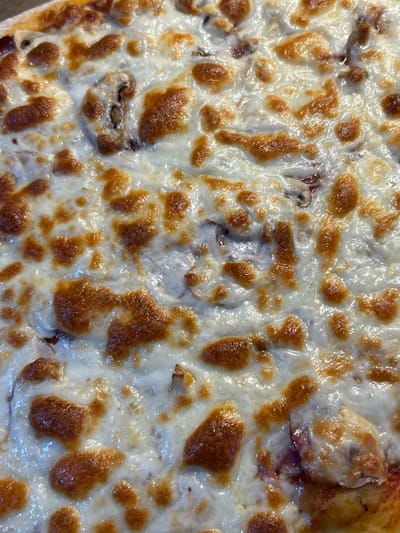 The image is a close-up, overhead shot of a pizza with melted cheese and visible toppings. The cheese is golden brown in spots, indicating it has been baked. Toppings that appear to be mushrooms and possibly some red sauce are visible beneath the cheese. The pizza is likely in a home or restaurant kitchen environment, and is not in the city of Shepelyovo, Russia. There are no people or actions depicted in the image.