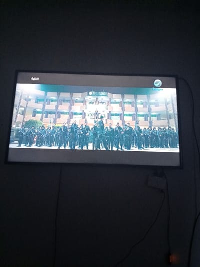 The image shows a television screen displaying a scene of many people wearing military uniforms standing in formation in front of a building. The people are facing the camera and are in a large group. The building appears to be a school or government building. The screen is mounted on a wall in a room with a dark grey wall. There are several electrical cords visible in the image. The location is near the entrance to the Alexandria University campus, in the city of Alexandria, Egypt.