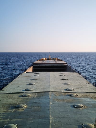 The image shows the deck of a cargo ship at sea. The deck is flat and gray, with several circular hatches visible. The ocean is blue and calm, with gentle waves. The sky is clear and pale blue. The ship appears to be moving forward, as indicated by the perspective. There are no people visible in the image. The location is stated to be the city of Prislon', Russia.