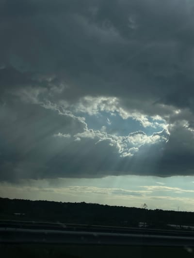 The image depicts a cloudy sky with sunbeams shining through a break in the clouds. The surrounding clouds are dark and ominous. A treeline is visible on the right side of the frame. The location is Georgi-Dobrevo, Bulgaria.