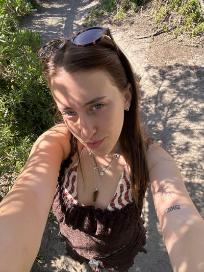 A young woman poses outdoors in bright sunlight. She wears sunglasses perched on her head, a brown ruffled top, and a patterned bikini top. A silver necklace with flower-shaped charms adorns her neck. A tattoo of the year "2002" is visible on her arm. She has a nose piercing and wears an earring. The backdrop includes greenery, ground, and shadows. The image is angled, creating a perspective looking down on the subject.