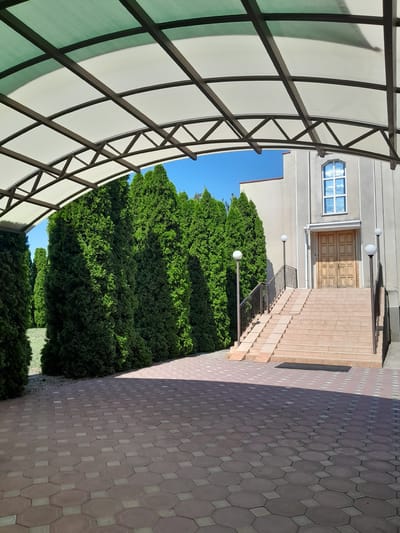 The image depicts the entrance of a building in the city of Illinka, Ukraine. A large, arched canopy with a translucent white roof supported by dark metal beams extends over a paved area. The paving consists of hexagonal, reddish-brown tiles with lighter, square accents.
To the left of the paved area, a row of tall, dense green cypress trees lines the property. Beyond the trees, a grassy area is visible.
To the right, a wide set of light-colored stone stairs leads up to the entrance of a building. The building's facade is a light gray color, with a prominent wooden double door at the top of the stairs. Above the door, a tall, arched window with multiple panes is visible. Two modern lampposts with spherical white lights stand on either side of the staircase. A ramp runs parallel to the left side of the stairs, providing an accessible route.
The sky is a clear, bright blue. The scene is captured from a low angle, looking towards the building entrance from under the canopy.