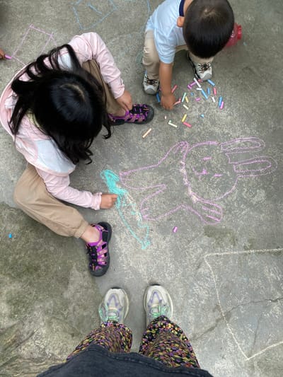 The image depicts an overhead view of two young children drawing with chalk on a concrete surface. A girl with long black hair, wearing a pink long-sleeved shirt and beige pants, is kneeling and drawing with a light blue chalk. She has on purple and black sandals. Beside her, a boy in a light blue shirt and beige pants is standing and picking up chalk sticks. He is wearing grey and white sneakers. Scattered around them are various colored chalk pieces, including pink, yellow, blue, and white. A pink chalk drawing of a rabbit-like creature is visible on the ground. The environment appears to be an outdoor area, likely a pavement or courtyard, with some cracks visible in the concrete. The overall scene suggests a playful activity by the children. The location is specified as Dujiangyan City, China.