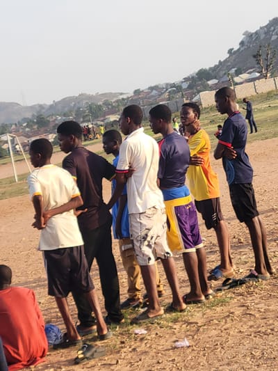 A row of seven Black African men, wearing various casual athletic clothing including shorts, appear to be performing some form of a bow or prayer on a dirt field. Behind the row is a slightly blurred view of a cityscape at the base of a rock outcropping. There is a red bag next to the last person in the row. The overall area appears to be on the outskirts of a developed area in Ibo, Nigeria.