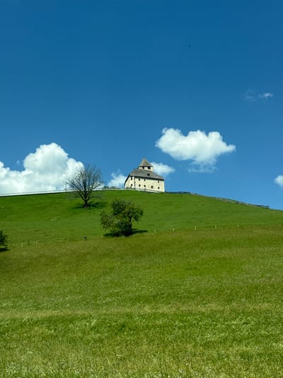 A medium shot captures a building situated atop a grassy hill under a clear blue sky with scattered white clouds. The building, appearing to be a church or a similar structure, has a light-colored facade with dark windows and a multi-layered roof, including a prominent tower. A leafless tree stands to the left of the building on the hill, and a smaller, leafy tree is positioned further down the slope. A wooden fence runs along the crest of the hill, encircling the building. The foreground consists of rolling green fields with a few scattered small plants or flowers. The image is taken from a lower vantage point, looking up at the hill and the building. The overall scene evokes a pastoral setting. This image is located in San Martin de Tor - San Martino in Badia - St. Martin in Thurn, Italy.