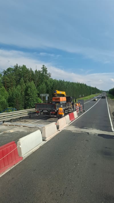 The image shows a highway construction or repair site. A black flatbed truck with a yellow crane is parked on the right side of the road, partially blocking one lane. The truck has an orange box-like attachment on its bed. Three construction workers wearing orange safety vests are standing near the truck and the road. The road is divided into lanes by white lines. Red and white traffic barriers are placed along the edge of the road. In the background, there is a dense forest of green trees and a blue sky with some white clouds. A white car and a truck are visible driving on the highway in the distance. The scene is set in Staryi Alzamai, Russia.
