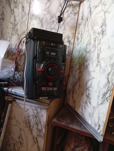 In Suklieia, Moldova, a black LG mini hi-fi system is placed on a shelf against a marbled wall. The system has a display showing "FM 89.60" and a CD player. Above the system, a black plastic device with wires connected to it is mounted. Several cables are visible around the audio system and extending to the left. The background wall has a white and gray marble pattern.