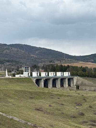 The image displays a concrete dam or spillway structure located within a rural landscape near Баня, Bulgaria. The central feature is a substantial grey concrete edifice, comprising six large, arched lower openings, likely conduits for water, situated above a flat base. Directly above these arches, six rectangular concrete structures with open tops are visible, potentially housing operating mechanisms for sluice gates. A thin, light-blue metal railing runs horizontally along the top edge of the main concrete structure.
To the left of the concrete structure, on an elevated grassy embankment, a tall, slender white monument or obelisk stands upright. Behind this monument, a multi-story, unfinished or dilapidated concrete building with exposed framework and missing window panes is visible. Further into the midground, additional smaller structures, including one with a reddish roof, are interspersed among sparse trees.
The foreground consists of a gently sloping grassy terrain, characterized by patches of dry, light-brown vegetation. A narrow concrete channel or ditch is present in the lower-left corner of the frame. The background is dominated by a series of undulating hills, extensively covered with bare deciduous trees and scattered evergreen foliage. Utility poles are discernible on some of the distant hilltops. The sky is uniformly overcast with light-grey clouds. No individuals or active subjects are present in the scene.
