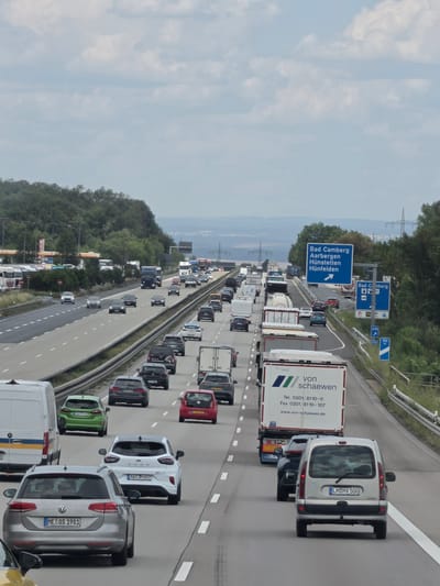The image depicts a heavily congested highway with multiple lanes of traffic in Germany, specifically on a section of the A3 autobahn heading towards Bad Camberg-Kernstadt. Numerous cars, trucks, and a few vans are stopped or moving very slowly. The sky is overcast with scattered clouds.

In the foreground, a silver Volkswagen Passat station wagon with license plate "ME OS 1981" is prominently visible. To its right, a white Ford Puma ST with license plate "AW JM 607" is also in frame. Behind these cars, other vehicles including a silver van with license plate "LM MX 500" are present.

Further down the highway, a large white truck with the branding "von schaewen" is stopped in traffic. Other trucks, some with trailers and others articulated, are also a significant part of the scene. The highway infrastructure includes guardrails and a concrete median.

On the right side of the highway, a blue highway sign indicates directions to "Bad Camberg", "Aarbergen", "Hünfelden", and "Hünfelden", with an arrow pointing to the right. Below this, smaller signs include an "M" for a McDonald's, a "U70" sign, and a "100 km/h" speed limit sign.

On the left side of the highway, a gas station with multiple pumps and convenience store is visible in the distance, along with a line of parked trucks. The surrounding landscape features a dense forest on a hill. Overhead, power lines and pylons are visible against the sky. The overall scene captures a typical traffic jam on a German autobahn, likely during peak hours or due to an incident.