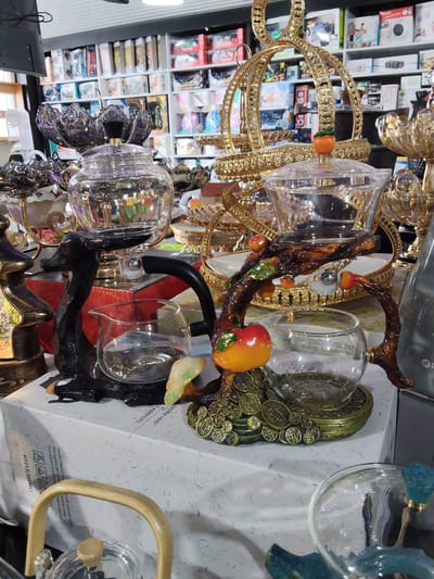 The image shows a display of decorative tea sets and serving dishes in a retail environment, likely a store. In the foreground, a black deer-shaped stand holds a glass teapot, and to its right, a branch-like structure with decorative apples holds another glass bowl. Behind these, ornate gold-colored tiered stands, possibly for serving fruits or desserts, are visible. The background consists of shelves stocked with various packaged goods. The lighting is artificial and appears to be from overhead fluorescent lights. The scene is static, with no people present. The overall impression is of a commercial setting showcasing elaborate decorative items.