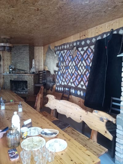 The image depicts the interior of a rustic wooden dining area, likely a restaurant or cafe, with a focus on traditional Caucasian decor. The ceiling is paneled with light-colored wood, and the walls are constructed from similar material. A large stone fireplace dominates the left side of the scene, with a dark opening and a decorative wooden sculpture above it.
On the wall behind the fireplace, a large woven tapestry with a geometric diamond pattern in shades of cream, pink, and blue hangs. To the right of the tapestry, a stuffed deer head is mounted, adding to the hunting lodge aesthetic. Two animal skins, one light-colored and fluffy and another darker and shaggier, are draped on the wall. Adjacent to these, a dark, heavy garment, possibly a traditional cloak, is hung.
The foreground is occupied by wooden tables, one in the foreground covered with a placemat, glasses, plates of food, and bottles. A bottle of milk, a glass jar with a golden lid, and a small cup with a cartoon character are visible. A smartphone with a colorful case lies on the table. Multiple clear glasses are also present.
The furniture includes long wooden benches with intricately carved sides, one resembling a dragon or mythical creature. Wooden chairs with dark seats are also scattered around. In the background, a wooden shelf unit with dark items is visible. The floor appears to be tiled. The overall impression is one of a cozy, traditional establishment. The presence of specific decorative elements and architectural style suggest the location could be Eltyubyu, Russia, a village known for its historical and cultural significance, often featuring such rustic architecture and traditional crafts. There are no people visible in the image.