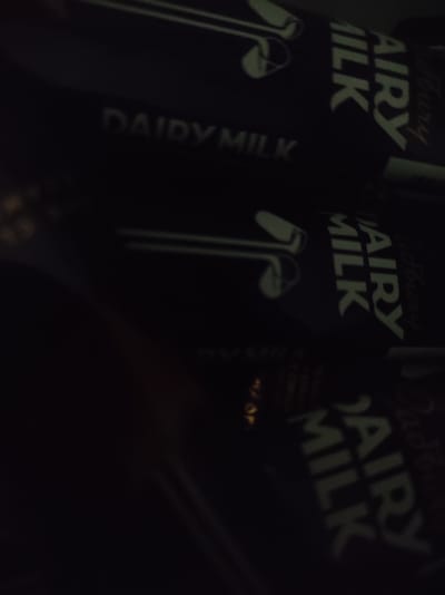 The image shows three Cadbury Dairy Milk chocolate bars, partially obscured by darkness and a blurred foreground. The bars are black with "DAIRY MILK" written in white lettering on each. The lettering is slightly angled, with the "DAIRY" appearing above and slightly to the left of the "MILK". The "AIRY MILK" is also visible on the middle and bottom bars, rotated vertically. The background is dark and indistinct, suggesting an indoor environment with low lighting. The location in Roranwala, Pakistan, is not visually evident from the image itself.