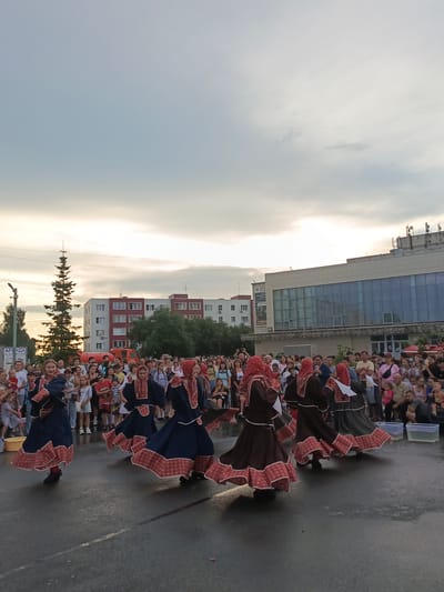 In this wide shot, a group of people are gathered outdoors, possibly for a festival or cultural event. The main subjects are several young women in traditional folk costumes, characterized by long, flowing dresses with wide skirts and decorative trim. They are dancing in a circular formation on a paved surface that appears slightly wet. The dancers are adorned in dresses of blue and brown, with red and white patterned borders on their hems and sleeves, and matching headscarves. They are facing away from the camera, their movements creating a sense of motion.
Behind the dancers, a diverse crowd of spectators is visible, many standing and observing the performance. The spectators are dressed in casual, modern clothing. In the background, there are multi-story buildings, one with a modern facade featuring large, reflective windows, and another with a more traditional brick design. A tall, slender evergreen tree stands to the left, adding to the outdoor setting. A fire truck is also partially visible among the buildings.
The time of day appears to be late afternoon or early evening, as indicated by the soft, diffused light and the overcast sky, with hints of sunlight breaking through the clouds. The atmosphere seems festive and engaging, with the audience attentively watching the performance. No specific emotions are overtly displayed by the crowd, but the scene suggests a lively community gathering.
Visible text includes a sign on a pole in the foreground on the left, partially obscured, but the letters "АРЕНДА" (ARENDA, meaning "rent" or "lease" in Russian) are discernible. A sign on the building in the background also displays some text and graphics, but it is too small to be clearly read. The overall setting suggests a public square or plaza in a town or city in Russia.