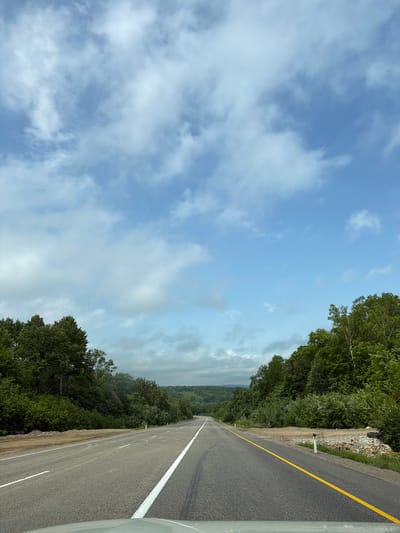 The image shows a two-lane asphalt road stretching into the distance through a landscape of rolling hills covered in dense green forest. The road has white dashed lines separating the lanes and a solid yellow line along the right edge. The sky above is a bright blue with scattered white cumulus clouds. The trees on either side of the road are lush and vibrant, indicating a temperate climate, likely summer. The road appears to be in good condition, with some minor debris visible on the shoulder. The perspective is from a vehicle traveling forward on the road. There are no people or any other discernible structures or objects in the immediate vicinity of the road, suggesting a rural or wilderness setting. This road is identified as being located in Teployozersk, Russia.