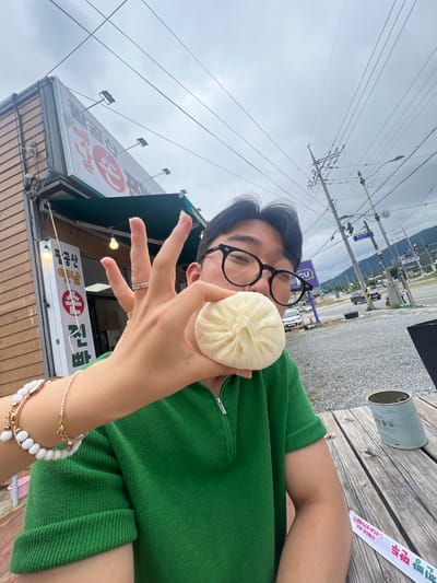 A person wearing a green zippered polo shirt and black-rimmed glasses holds a white steamed bun to their face in front of a building with a wooden exterior and a green awning. The building has a sign with Korean characters. The person is seated at a wooden table outdoors, with a wooded area and a street visible in the background. The sky is overcast.