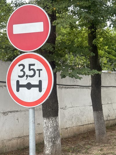 In Dedovsk, Russia, two traffic signs are mounted on a single metal pole. The upper sign is a red circle with a white horizontal bar, indicating "Do Not Enter." Below it, a circular sign with a red border displays "3.5T" and a graphic representation of an axle with a load limit. Both signs are positioned in front of a concrete wall and two trees with white-painted bases. The background features dense green foliage.
