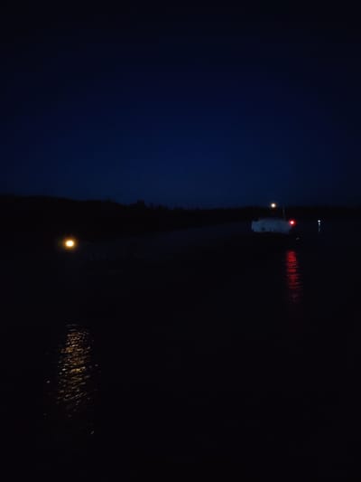 At night in Badozhskoi Pogost, Russia, a body of water reflects light from visible structures and points of light. In the middle ground, a white structure with a bright light on top and a red light on the side stands on what appears to be land. To the left, a single orange light is reflected in the water. Trees are visible on the horizon.