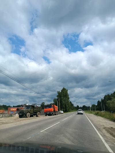 The image depicts a roadside scene in Снопок Старый, Russia. A paved road with a white line marking the lane runs horizontally across the frame. On the left side of the road, there are two parked trucks. The first truck is a green military-style truck with a flatbed, and the second is an orange dump truck loaded with gravel. Adjacent to the trucks is a construction material store with a sign reading "ЦЕМЕНТ" (Cement). In the background, a line of trees and power lines are visible against a cloudy sky. Further down the road, a white car is driving away from the viewer. The scene appears to be an overcast day.