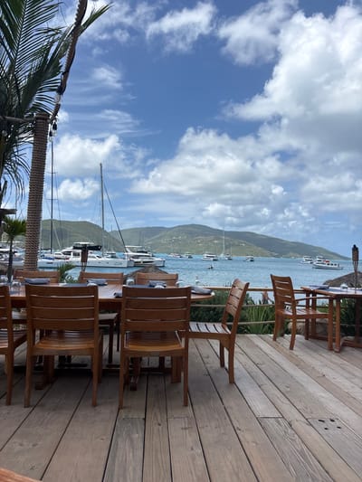 The scene depicts an outdoor dining area on a wooden deck overlooking a marina in Spanish Town, British Virgin Islands. In the immediate foreground, a weathered light brown wooden deck occupies the lower half of the image. A large rectangular wooden dining table is positioned centrally, set with blue placemats or napkins and upturned clear glasses. It is surrounded by at least six matching wooden chairs with slatted backs. To the far right, a smaller square wooden table with two similar chairs is visible.
A thick rope-wrapped column, possibly part of a palm tree trunk, is on the far left, from which string lights with exposed bulbs are strung. Green palm fronds extend from this column at the top left. A low rope railing and dense green foliage separate the deck from the water directly beyond.
The midground features a calm, turquoise body of water, identified as a marina. Numerous boats are moored or anchored, including several white sailing yachts, both monohulls and catamarans. One prominent white catamaran displays "Dream Yacht Charter" on its hull. Smaller motorboats and dinghies are also present. A dark-colored inflatable boat with at least one individual standing or sitting within it is visible in the middle of the bay.
The background consists of verdant, hilly landmasses under a clear blue sky interspersed with scattered white cumulus clouds. No individuals are seated at the foreground tables. A tiki torch with a thatched top is visible near the smaller table on the far right.
