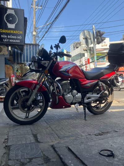 A red Suzuki motorcycle is parked on a grey, patterned tiled sidewalk in Hòa Cường Ward, Vietnam, under a clear blue sky. The motorcycle, oriented facing left, rests on its kickstand. It features a transparent front windshield, two circular headlights with chrome rims, two rearview mirrors, and a black storage case mounted on the rear rack. The front wheel has a disc brake, and both wheels display multi-spoke alloy rims. A small white plastic bag is attached near the engine. In the lower right foreground, a dark, C-shaped metal ring lies on the pavement. To the left, a dark storefront sign for "DANAWATCH" with a white watch icon is visible, along with Vietnamese text "SỬA ĐỒNG HỒ LẤY NHANH" (Watch Repair Get Fast). The background includes a dense network of utility wires on a pole, a traffic light, and various urban buildings, one bearing the partial text "ank Live." A white vehicle is partially visible on the street to the far right. No individuals are present in the brightly lit scene.