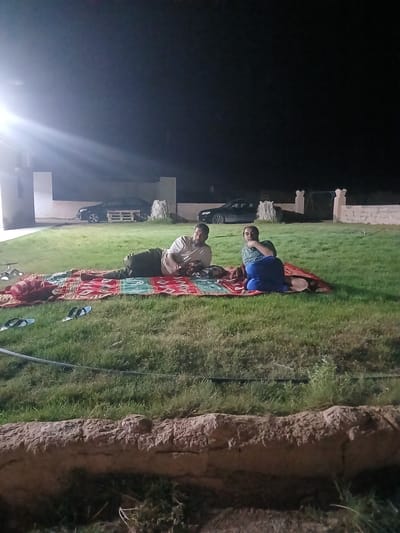 In the city of Hun, Libya, on a dimly lit grassy area at night, three people are casually lying on a patterned mat. A man in a light-colored t-shirt and dark pants is reclined on his left side, facing to his right. Beside him, a person in a blue t-shirt and blue pants is sitting up, leaning on their right arm and looking to their left. Another individual, dressed in a light-colored shirt and dark pants, is partially visible on the far left. In the background, two cars are parked, and architectural structures are dimly illuminated. The foreground features a textured, rocky border.