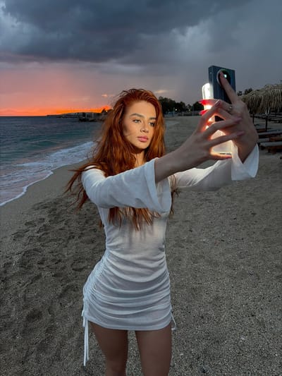 A photograph taken on a beach in Alimos, Greece shows a woman with long red hair, wearing a white mini dress, holding a smartphone with a light attachment. The woman is facing the camera, and the phone is positioned in front of her. The background features a sunset over the sea with a cloudy sky to the left. The beach is sandy, and there are waves near the shore. A thatched roof structure is visible in the upper right corner.