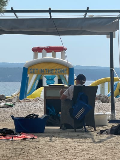 This is a vertically oriented, full shot of a beach scene in Crikvenica, Croatia. The foreground is dominated by a sandy beach and a stone wall on the right. In the center, a person wearing a black baseball cap and a light-colored t-shirt is seated in a dark-colored woven chair, facing away from the camera. A blue backpack with white stripes is placed on the chair behind them.
Behind the seated person, a large, inflatable water slide structure is visible, with segments in yellow, blue, and white. The words "GET IN THE ZONE" are printed vertically in yellow on a blue banner on the slide. A red cylindrical object with the word "LIFEGUARD" printed on it in white is also part of the inflatable structure.
The background shows a body of water and a hazy, distant coastline with rolling hills. The sky is clear and bright, suggesting it is daytime, likely late morning or early afternoon, with good weather. A dark fabric canopy is visible in the upper left corner. On the ground to the right, a blue plastic bin holds some items, and a pink and orange life vest is draped over it. The overall impression is a recreational area at a seaside resort.
