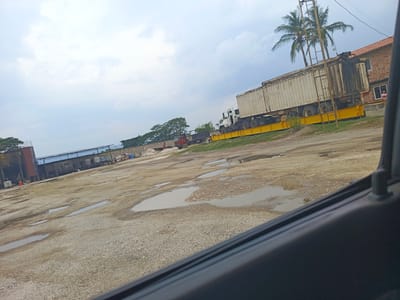 The scene depicts an industrial yard in El Bajío, Venezuela. The ground is gravel and dirt with numerous puddles. A semi-truck with a large white trailer is parked on a yellow platform scale. Behind the truck is a two-story red brick building with a tiled roof and several palm trees. In the background, there are other industrial structures, including what appears to be a tank and a covered area. A red truck is visible in the distance. The sky is overcast. The photo is taken from inside a vehicle, with a portion of the vehicle's interior visible in the lower right corner.