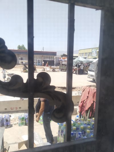 This is an outdoor, eye-level shot of a busy marketplace in Danghara, Tajikistan, viewed through a metal grate. The scene is bustling with activity, likely during the daytime given the bright sunlight and shadows. In the foreground, a metal grate with ornate scrollwork partially obstructs the view. Several bottles of water, shrink-wrapped in plastic, are stacked on the ground to the left and right.
The middle ground reveals a street or open area with various market stalls and vendors. One stall, covered by a patterned green awning, displays a large quantity of what appear to be melons or similar round produce. Several people are visible around this stall, engaged in apparent transactions or preparing goods. To the left, a man is pushing a cart, and a small cart with wheels is parked nearby. Further back, a modern building with glass windows and an orange trim can be seen, possibly a shop or administrative building. Another yellow building with signs is visible in the distance. Various vehicles, including a white van and other carts, are present, suggesting active commerce.
The background shows a clear sky with distant hills or mountains. The ground is a mixture of dusty earth and pavement. The overall impression is one of a vibrant, functional market in a Central Asian city.