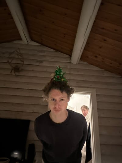 A man with curly hair and a tiny Christmas tree on his head peers into the camera. He wears a black shirt and stands inside a room with light-colored wood paneling. In the doorway behind him, another person’s face is visible. A rustic wooden ceiling and beam are seen on the left. In the bottom right, there is a glimpse of a dark television set.