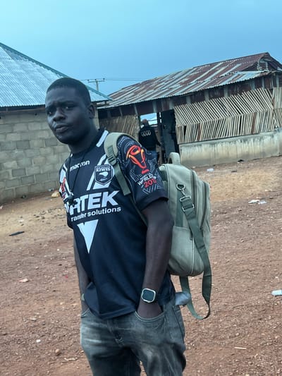 The image shows a young Black man wearing a black sports jersey with white and red accents, a green backpack, and gray jeans. He is standing in an outdoor environment with buildings in the background. The buildings appear to be constructed with corrugated metal roofs and brick or wood walls, suggesting a rural or semi-urban setting. The ground is covered in dirt and gravel. The man is looking towards the camera with a neutral expression. The sky is overcast. There is a person in the background wearing a jacket with the word "GOOD" on the back. The location is identified as Durimi, Nigeria.
