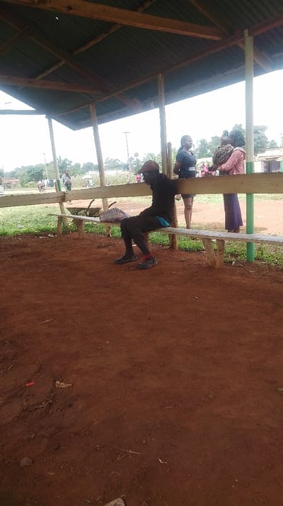 The image captures a scene within an open-sided, covered structure in Siaya Township ward, Kenya. The ground within the structure consists of reddish-brown dirt with scattered small debris. The roof is corrugated metal, supported by wooden beams and posts, some painted green. Wooden benches are integrated into the structure's perimeter.
In the left foreground, a man sits on a wooden bench, facing right. He wears a dark long-sleeved top, dark trousers, dark shoes with red accents, and a reddish-brown cap. A blue garment is visible at his waist. A multi-colored patterned bag rests on the bench to his left.
In the middle ground, positioned behind the benches, two women stand. The woman on the left stands facing left, wearing a dark short-sleeved top and dark lower garments. To her right, a second woman stands facing left, wearing a light-colored long-sleeved top and a long purple skirt, carrying an infant on her back secured by a patterned fabric wrap.
The background reveals an expanse of reddish-brown dirt, green grass, and trees. A wheelbarrow with contents is visible behind the benches. Further back, additional blurred figures, utility poles, and distant buildings are discernible under an overcast sky.