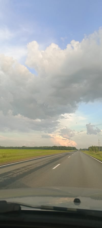 The image shows a view from inside a car driving on a two-lane road. The road stretches into the distance, bordered by a bright green field on the left and a line of trees on the right. The sky is filled with dramatic clouds, with patches of blue sky visible. The lighting suggests it is late afternoon or early evening, with the sun casting long shadows on the road. The overall mood is serene and expansive, evoking a sense of journey and open road. No people or text are visible in the image.