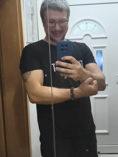 A man with short, grey hair and glasses smiles while taking a mirror selfie. He is wearing a black t-shirt and has a tattoo on his left bicep. He is also wearing two bracelets on his right wrist. In the background, a white door with a arched window is visible. The image appears to have been taken indoors, possibly in a residential setting. There is no information in the image to suggest it is located in Eitorf, Germany.