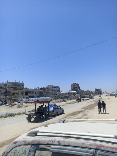 This is a wide shot of a dusty road in Gaza, with buildings under construction and some damaged structures in the background. The sky is clear and blue. In the foreground, a white vehicle's roof is visible, covered in dust. Further down the road, a horse-drawn cart is transporting several people and some cargo. Two men are walking on the side of the road, one of them carrying a child. Several other vehicles and makeshift structures are scattered along the road, suggesting a busy, possibly informal, marketplace or residential area. The overall impression is one of ongoing development and potentially challenging living conditions.