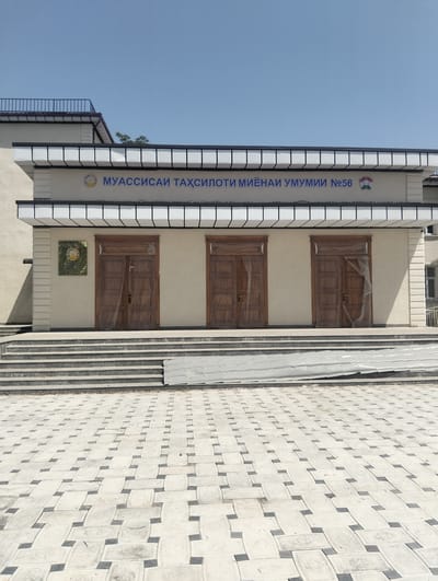 The image depicts the facade of a public building, identified by the signage as "МУАССИСАИ ТАҲСИЛОТИ МИЁНАИ УМУМИИ №56" which translates to "General Secondary Education Institution No. 56" in the city of Danghara, Tajikistan. The building has a light-colored facade with three prominent wooden double doors at the entrance. These doors are covered with transparent plastic sheeting. Above the doors, a decorative white cornice with dark trim runs across the building. A plaque with an emblem is mounted on the wall to the left of the central door.
The entrance is accessed via a wide set of concrete stairs. The ground in front of the stairs and extending outwards is paved with a patterned interlocking tile design in shades of light gray and dark gray. A large sheet of corrugated metal or plastic sheeting is laid out on the ground just in front of the bottom of the stairs, appearing to be construction material or protective covering.
The sky above is clear and blue, indicating a sunny day. The building's architecture appears modern, with clean lines and minimalist decorative elements. The overall impression is of an institutional building, possibly a school, in an urban or suburban setting in Tajikistan.