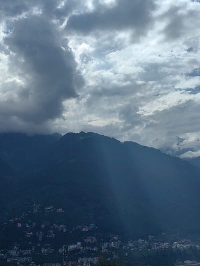 The image captures a panoramic view of Siumsa, India, under a dramatic sky filled with heavy, dark clouds and patches of bright sunlight. The foreground is dominated by the lush, dark green slopes of a mountain range. A dense, scattered settlement is visible on the lower slopes of the mountain, with buildings of varying sizes and colors nestled amongst the trees. The architecture appears to be a mix of residential and commercial structures. In the distance, higher mountain peaks are shrouded in mist and clouds, with one peak showing a hint of snow. The lighting suggests it could be late afternoon or early evening, with shafts of sunlight breaking through the clouds, casting rays onto the landscape. The overall atmosphere is one of natural grandeur and human habitation coexisting in a mountainous region.