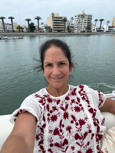 An outdoor, eye-level, medium shot shows a woman taking a selfie from a boat on the water. The woman is in the foreground, her face is in focus, and she is smiling. She has dark hair pulled back from her face and is wearing a white blouse with red floral embroidery. Behind her, the water is choppy, and a cityscape lines the horizon. The city appears to be La Punta, Peru, with buildings and palm trees visible. Several small boats are anchored in the water. The sky is overcast.