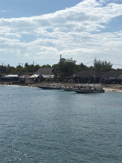 In Kampung Toyapakeh, Indonesia, the image captures a bright, sunny day with a blue sky filled with fluffy white clouds. The foreground shows the ocean with gentle waves reflecting the sunlight. Along the shoreline, a rocky beach separates the water from a line of low-rise buildings and some trees. Several boats are visible near the shore. One is a white boat with an orange stripe and the words "OCEAN BIKER CRUISE" visible on its side, carrying passengers. Another boat is partially visible to the left. On the shore, a group of people dressed in white are gathered, some holding umbrellas. There are also some smaller structures with awnings. Power lines and a cell tower are visible among the trees behind the buildings.