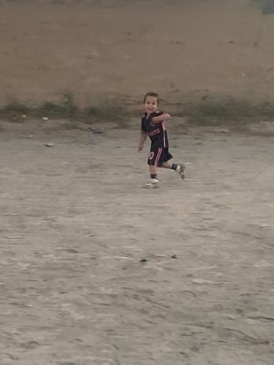 A young boy with short brown hair, wearing a dark soccer jersey with pink accents and the number "10" on his shorts, runs across a dirt field. The jersey has the text "YBTO" visible on it. He is looking towards the camera with a smile. The background consists of a muted brown wall or embankment. The ground is a dusty, gray-brown surface with some scattered debris, including small white objects and what appears to be a pink piece of paper. The lighting suggests it might be late afternoon or early morning. The scene is likely in a less developed area, possibly a park or a casual sports field. The location is in Amondara, Tajikistan.